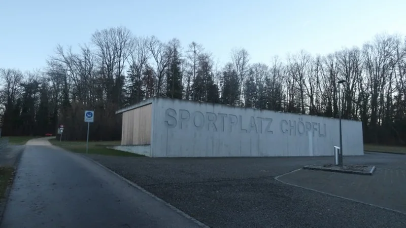 Das Garderobengebäude des Sportplatz Chöpfli in Hofstetten-Flüh, mit dem Namen an der Betonwand und Bäumen dahinter